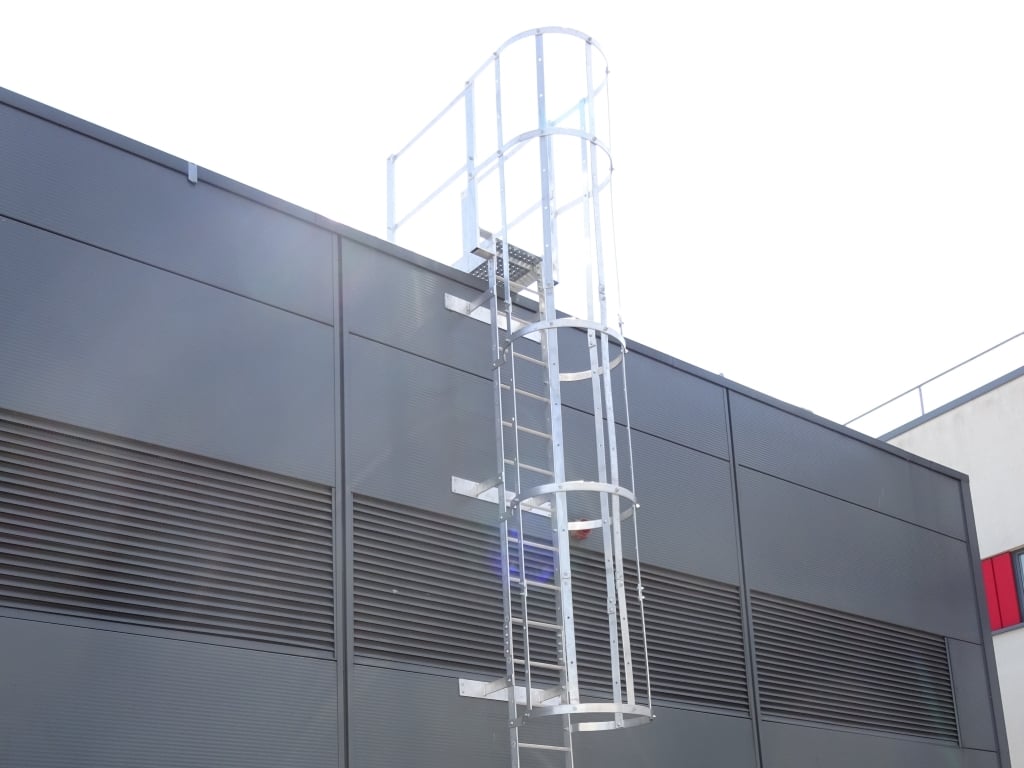 Aluminium fixed ladder with hoops going up the side of a building onto an industrial roof.